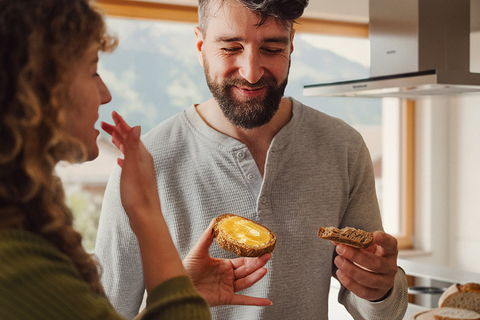 Zwei Personen in moderner Küche, eine hält ein belegtes Brötchen, im Hintergrund Kücheninsel mit Brot und Espressokanne