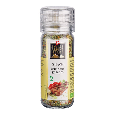 Transparente Gewürzmühle mit durchsichtigem Deckel. Auf dem Etikett steht „Grill-Mix / Mix pour grillades“ unter dem Swiss Alpine Herbs Logo. Im Glas sind grobe Kräuter, Gewürze und Salzkristalle sichtbar. Unten sind ein gegrilltes Fleischstück, rote Peperoni sowie das Bio-Logo abgebildet.
