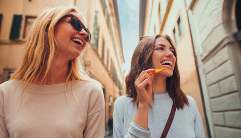Zwei Frauen gehen in einer engen Gasse zwischen hohen Gebäuden, eine isst getrocknetes Mango Stück