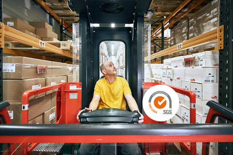 Person in gelbem T-Shirt steuert Gabelstapler in Lager mit Regalen voller Kartons