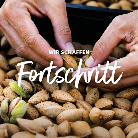 Hände öffnen eine Schale mit Mandeln auf einem Haufen Mandeln mit dem Text 'WIR SCHAFFEN Fortschritt'
