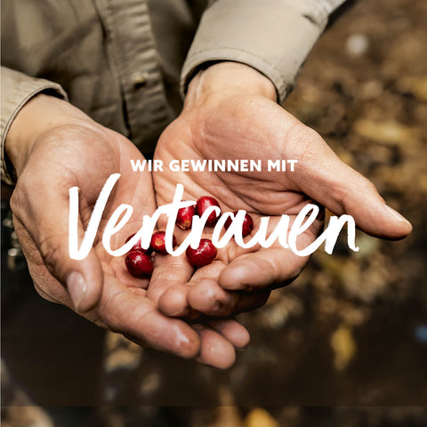 Zwei geöffnete Hände halten mehrere kleine rote Beeren vor unscharfem Hintergrund mit dem Text 'WIR GEWINNEN MIT Vertrauen'